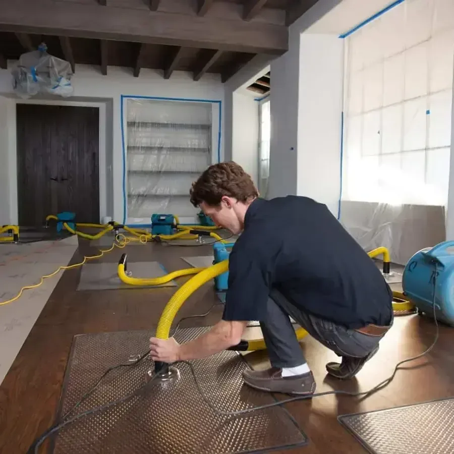 A technician kneeling over a hardwood floor with drying mats and yellow hoses attached